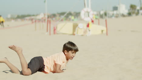 Front-view-of-excited-kid-kicking-ball-and-falling-on-sand